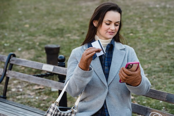 Mulher sentada em um banco com o celular e um cartão de banco em mãos.