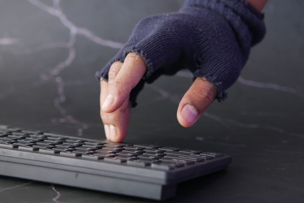 Imagem simulando um ataque hacker, na qual podemos ver uma mão humano usando uma luva escura até a metade dos dedos e digitando em um teclado de computador.