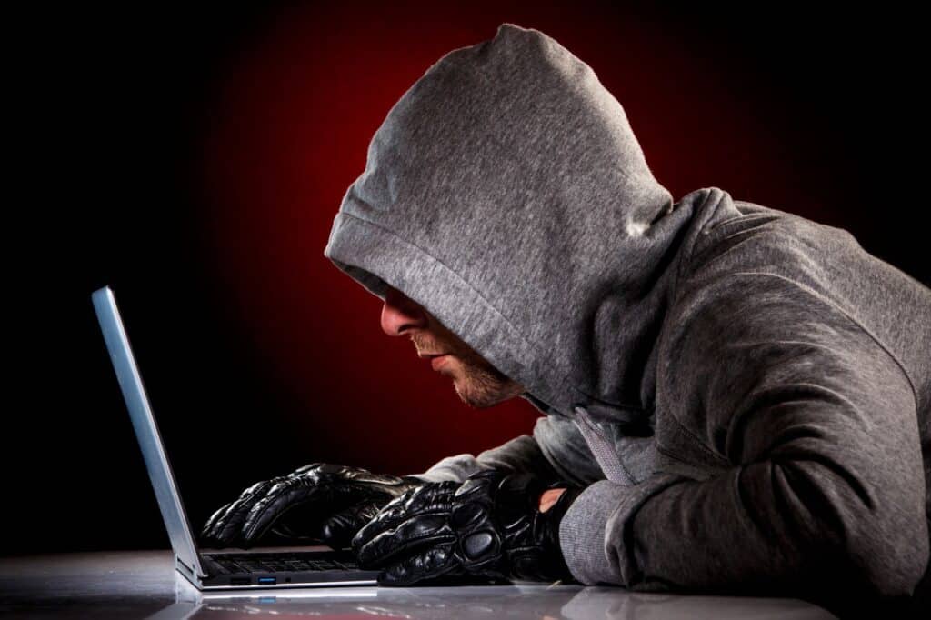 Imagem representativa de ataque hacker, na qual vemos uma pessoa de capuz, roupas escuras, luvas e um fundo vermelho, utilizando um computador com o rosto muito próximo à tela.